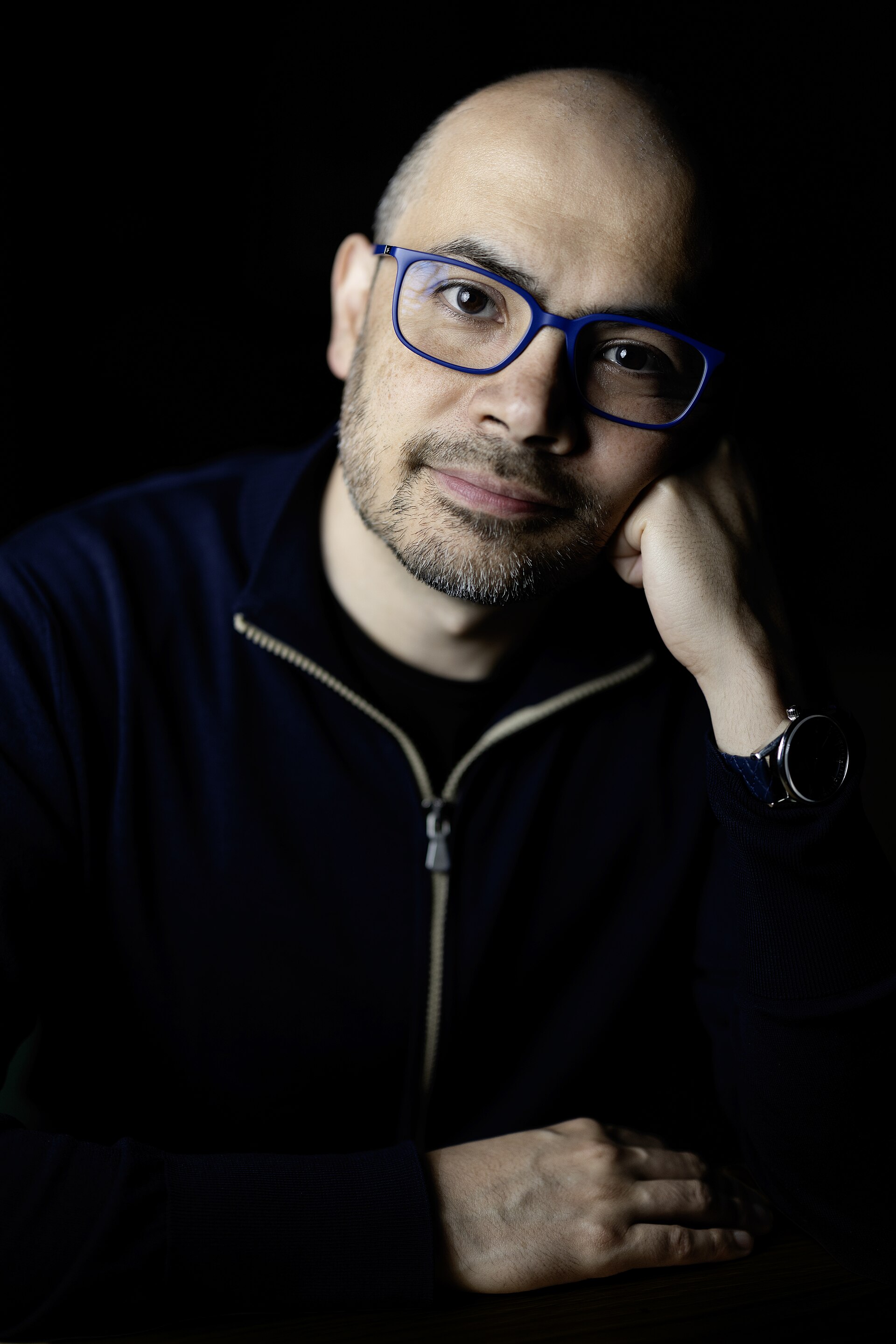 Demis Hassabis seated against a dark background, wearing blue-framed glasses